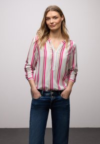 Femme aux longs cheveux blonds portant un chemisier rayé à manches longues rose, blanc et beige, les mains dans les poches d'un jean bleu foncé.