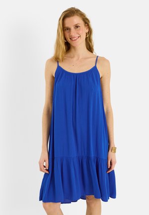 Femme souriante portant une robe bleue ample sans manches avec des fines bretelles et un ourlet volants, debout devant un fond clair uni.