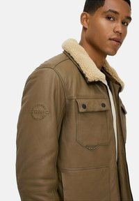 Braune Lederjacke mit einem Lammfellkragen, mehreren Vorderaschen und einer strukturierten Oberfläche. Mit geprägtem Detail am Ärmel.