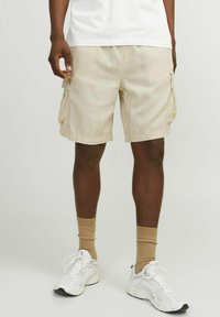 Shorts cargo beige avec poches latérales, fabriqués à partir d'un matériau léger. Portés avec des baskets blanches et des chaussettes crew tan. Coupe décontractée.