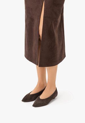Kvinde iført mørkebrun sko med spids tå og en matchende brun nederdel med en slids foran, der viser hendes underben.