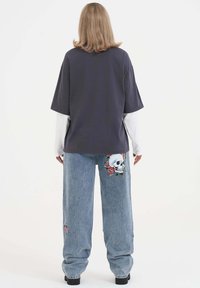 Overmaatse donkergrijze t-shirt gelaagd over een wit longsleeve shirt, gecombineerd met lichtblauwe denim jeans met borduursel van een schedel en roos.