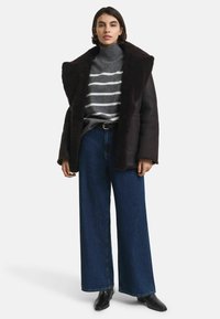 Veste en shearling marron foncé avec un large col, col roulé rayé gris, et jeans bleus à jambes larges. Des bottes noires à la cheville complètent la tenue.