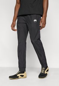 Μαύρο ελαφρύ παντελόνι με ελαστικές μανσέτες και διακριτικό λογότυπο στο μπροστινό μέρος, σε συνδυασμό με μαύρα και μπεζ παπούτσια από δίχτυ.