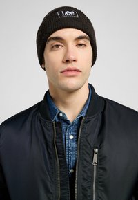 Berretto lavorato a maglia nero con logo "Lee" ricamato in bianco. Indossato sopra una camicia di jeans e una giacca bomber nera, mostrando un look casual a strati.