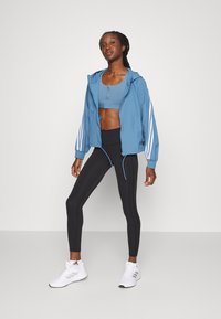 Casaco azul claro com fecho éclair e riscas brancas nas mangas, combinado com um soutien desportivo a condizer,legging pretos e sapatilhas atléticas brancas.