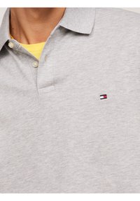 Polo gris en tissu doux. Il présente un col classique, deux boutons et un petit patch de logo en rouge, blanc et bleu sur la poitrine.