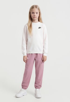 Lyshåret pige iført hvid Nike-sweatshirt, lyserøde cargobukser og hvide sneakers, stående mod en ensfarvet lys baggrund.
