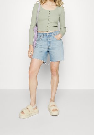 Vrouw draagt een lichtgroen kort knooptopje, lichtblauwe spijkershorts, pluizige beige sandalen en heeft een lavendelgekleurde gebreide schoudertas bij zich.