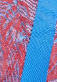 Tejido texturizado en rojo con patrones abstractos en azul, junto a una cinta azul plana y estriada. Los materiales parecen suaves y tejidos.