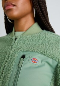 Green bomber jacket with textured fleece and smooth fabric. Features a zip pocket and a circular "Dickies" logo patch on the chest.