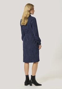 Robe en suède bleu marine avec une taille ceinturée, des manches longues et une coupe à mi-genoux, accompagnée de bottines à talons noires.