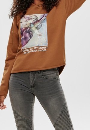 Bruine hoodie met een engel-graphic en tekst "Ik geef al mijn geld uit aan design schoenen", gecombineerd met grijze aansluitende jeans met geribbelde knie-details.