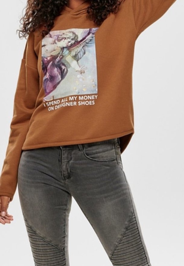 Sweat à capuche marron avec un graphisme d'ange et le texte « Je dépense tout mon argent en chaussures de créateurs », associé à un jean gris ajusté avec des détails côtelés aux genoux.