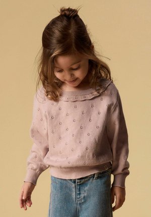 Noorpruunis juustes noor tüdruk, kellel on pealepandud juuksekoht, kannab roosat mustrilist kampsunit ja siniseid teksaseid, vaadates allapoole kerges naeratuses.