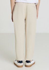 Beige, locker sitzende Hose mit strukturierter Oberfläche, elastischem Bund und einer einzigen Gesäßtasche, kombiniert mit weißen Sneakers und marineblauen Akzenten.