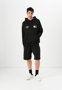 Sudadera con capucha negra que cuenta con un bolsillo frontal y gráficos, combinada con pantalones cortos negros. Zapatillas blancas y calcetines negros completan el conjunto.