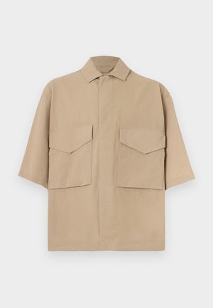 Camicia beige a maniche corte con due grandi tasche anteriori con patta e colletto classico, esposta su uno sfondo neutro.