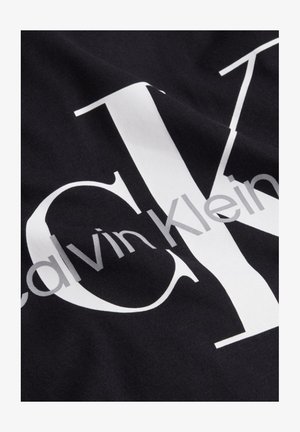 Čierna látka s veľkými bielymi písmenami "CK" a menším sivým textom "Calvin Klein" potlačeným diagonálne cez látku.