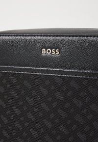 Juodas tekstūruotas maišas su lygaus odos viršumi, auksiniu "BOSS" logotipu ir subtiliu raštu pagrindinėje dalyje.