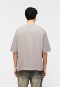 Άτομο που κοιτάζει προς τα πίσω, φοράει άνετο γκρι κοντομάνικο μπλουζάκι και ξεβαμμένα γκρι τζιν, πάνω σε απλό φωτεινό φόντο.