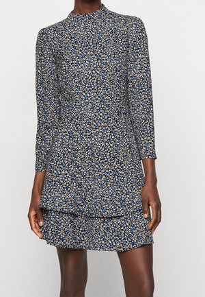 Femme portant une robe à manches longues à imprimé floral noir, bleu et blanc avec un col montant et une jupe à volants, debout devant un fond uni.