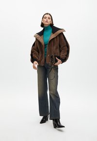 Brown faux fur jacket with black accents, green turtleneck, embellished dark denim jeans with gold studs, and black heeled boots.