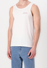 Hombre vistiendo una camiseta sin mangas blanca de Tommy Jeans y jeans de denim de color azul claro, de pie con los brazos relajados a los lados.