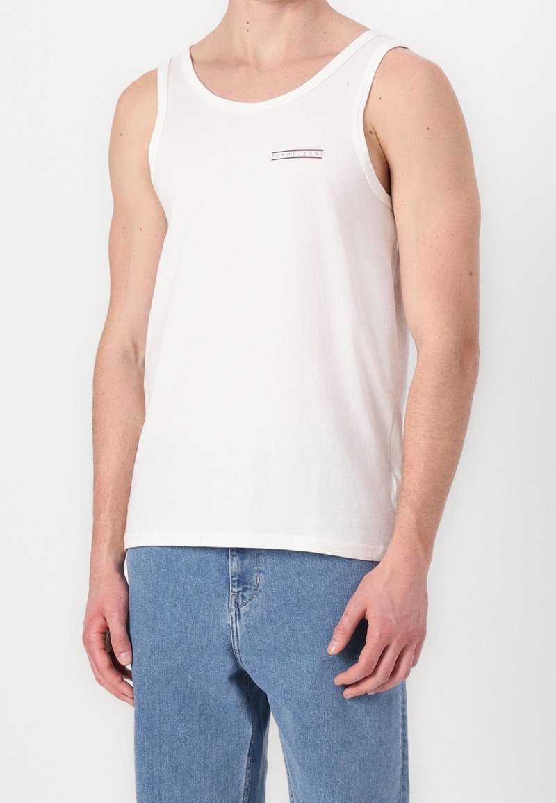 Hombre vistiendo una camiseta sin mangas blanca de Tommy Jeans y jeans de denim de color azul claro, de pie con los brazos relajados a los lados.