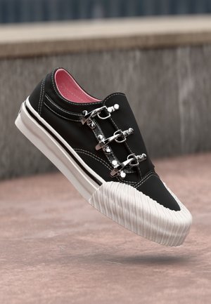 Czarny sneaker z białą gumową podeszwą i noskiem, wyposażony w trzy srebrne metalowe klamry oraz różową wyściółkę wewnętrzną, pokazany w powietrzu nad chodnikiem.