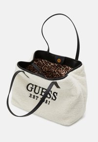Sac fourre-tout en polaire blanche avec des poignées en cuir noir, doté d'un intérieur à motif léopard et de l'inscription "GUESS EST 81" brodée en noir.