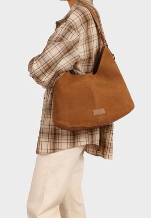 Persona con camicia a quadri beige e pantaloni crema che porta una grande borsa a tracolla in pelle scamosciata marrone sul braccio destro.