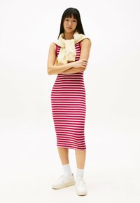 Donna in piedi con le braccia incrociate, che indossa un vestito midi senza maniche a righe rosse e bianche, sneakers bianche, calze e un maglione beige appoggiato sulle spalle.