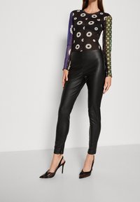 Leggings in pelle nera abbinati a un top trasparente nero con motivi floreali bianchi e maniche lunghe colorate. Tacchi alti neri a punta.