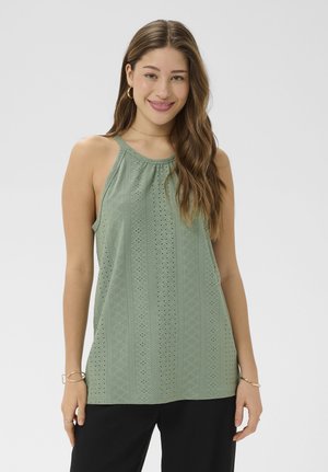 Žena s dlhými vlnitými vlasmi, oblečená v zelenom bezrukávovom topánke s dierkami a čiernych nohaviciach, usmievajúca sa, s pozlátenými kruhovými náušnicami a náramkami.