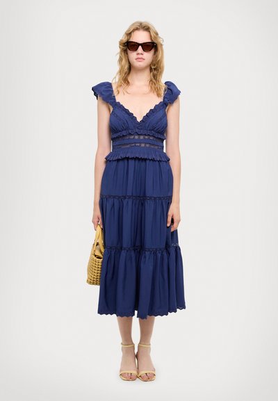 Navyblaues, gestuftes Maxikleid mit Rüschensäumen, Spitzenverzierungen an der Taille und lockerer Passform. Das Model hält eine gelbe, geflochtene Tasche.