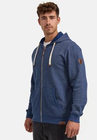 Sudadera azul con cremallera fabricada en tejido texturizado, que cuenta con capucha con cordón, dos bolsillos frontales y un parche de logo en la manga.