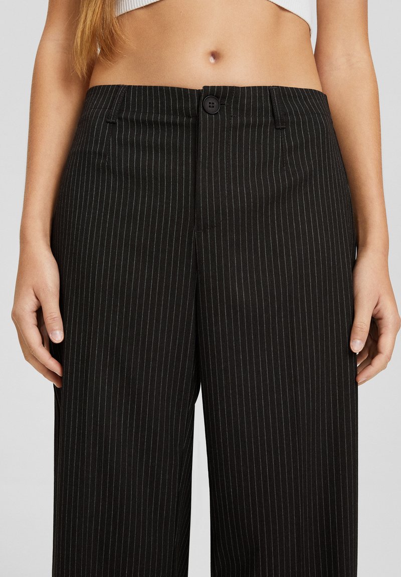 Pantalons à rayures noires avec taille haute, dotés d'une fermeture à bouton et de poches latérales, fabriqués à partir d'un tissu lisse.