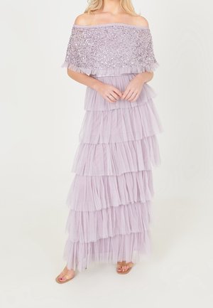 Kobieta w lawendowej sukni z odkrytymi ramionami, z cekinowym gorsetem i warstwową falbaną na spódnicy, stojąca boso na jednolitym tle.