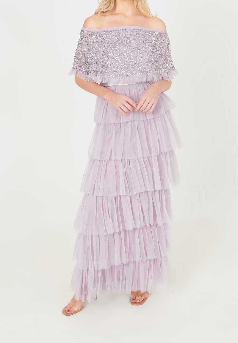 Femme portant une robe lavande à épaules dénudées avec un corsage orné de sequins et une jupe à volants superposés, debout pieds nus devant un fond uni.