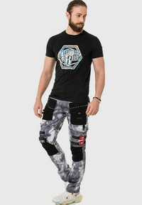 T-shirt noir avec un design graphique coloré. Pantalons cargo tie-dye gris avec plusieurs poches, accents noirs et quincaillerie décorative.