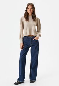 Beige geripptes Langarm-Crop-Top mit gerafften Schultern, kombiniert mit dunkelblauen weiten Jeans. Die Jeans haben ausgefranste Säume und einen lockeren Schnitt.