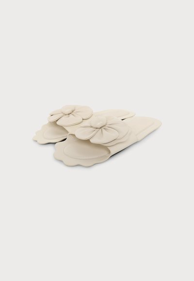 Sandales beige à enfiler avec bords festonnés et grandes décorations florales rembourrées sur les brides, présentées sur fond blanc.