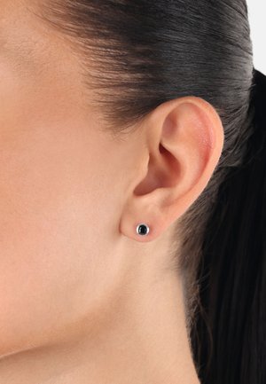 Pendiente de stud negro con forma redonda y una montura de tono plateado. El pendiente está colocado en la oreja de un modelo, destacando su diseño minimalista.