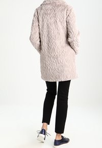 Puffig, ljusrosa kappa med en texturerad, vågig mönstring; bakvyn visar en krage och neddragna axelsömmar, ihopparad med svarta byxor och skor.