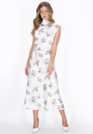 Donna che indossa un vestito bianco senza maniche con motivi floreali grigi, in piedi con una gamba leggermente avanti e tacchi alti bianchi.