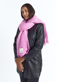 Vrouw draagt een zwart leren jasje en een grote roze sjaal met een wit label, staand met handen losjes gesloten tegen een witte achtergrond.