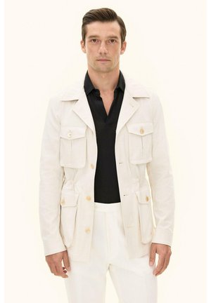 Man draagt een crèmekleurig jasje met grote voorzakken over een zwart overhemd met kraag en witte broek, staand tegen een lichte achtergrond.