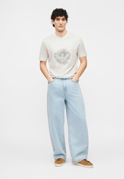 Νεαρός με σγουρά μαλλιά που φοράει λευκό t-shirt με σχέδιο, ανοιχτό μπλε τζιν παντελόνι φαρδύ και καφέ σουέντ αθλητικά παπούτσια, στέκεται με τα χέρια στις τσέπες.