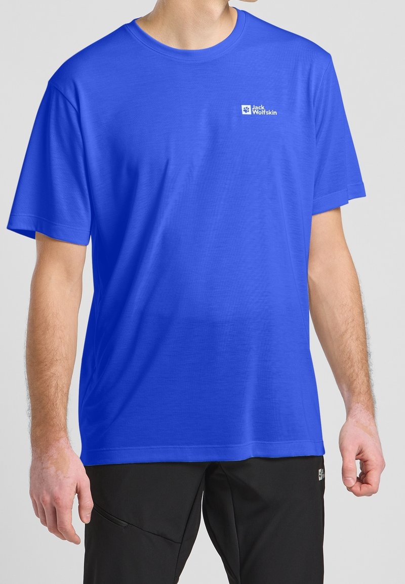 Homme portant un t-shirt à manches courtes bleu vif avec le logo "Jack Wolfskin" sur la poitrine et un pantalon noir, sur un fond uni.
